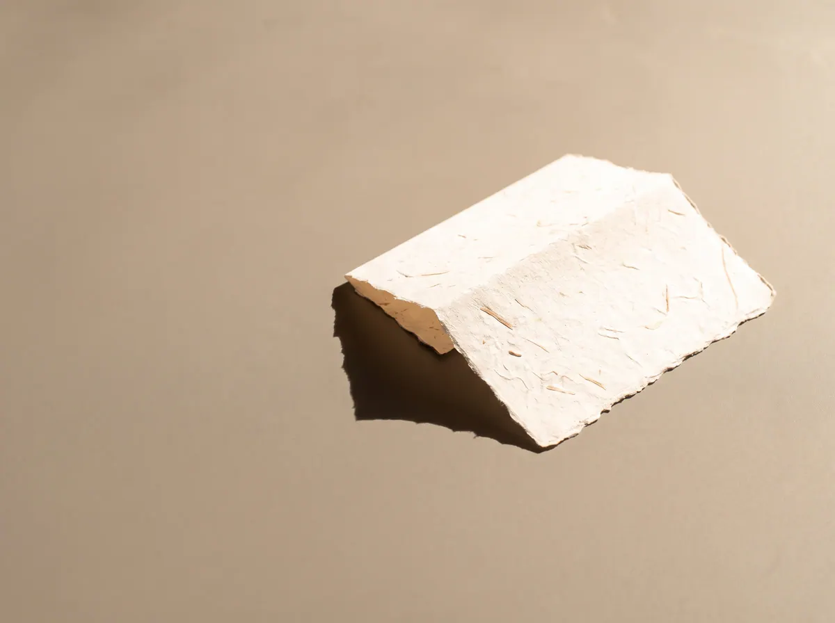 Une épaisse feuille de papier fait main, posée de biais sur une table, sous une lumière d'en haut.